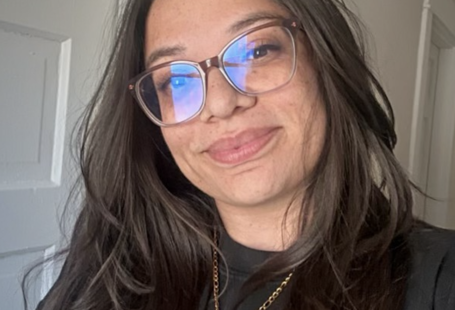 A smiling person with long, dark hair, wearing glasses and a black shirt, stands near an open door.